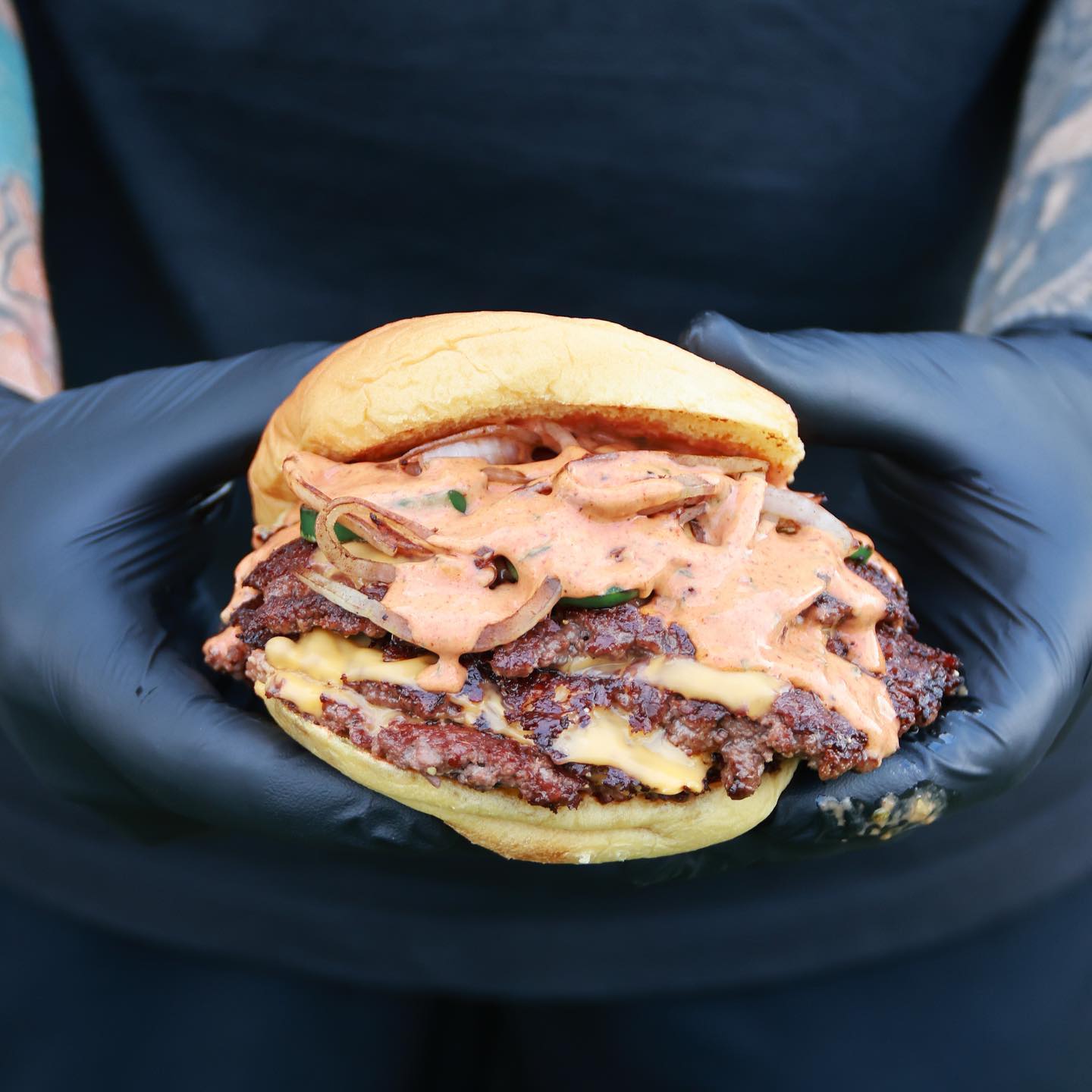 Smashed grass-fed burger with melted cheese, grilled onions, and signature sauce held in a black-gloved hand